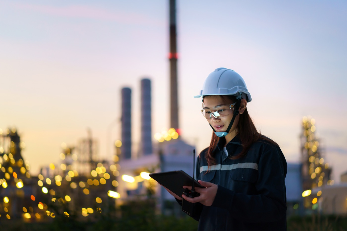 Engineer in a hard hat with a tablet at a refinery at dusk